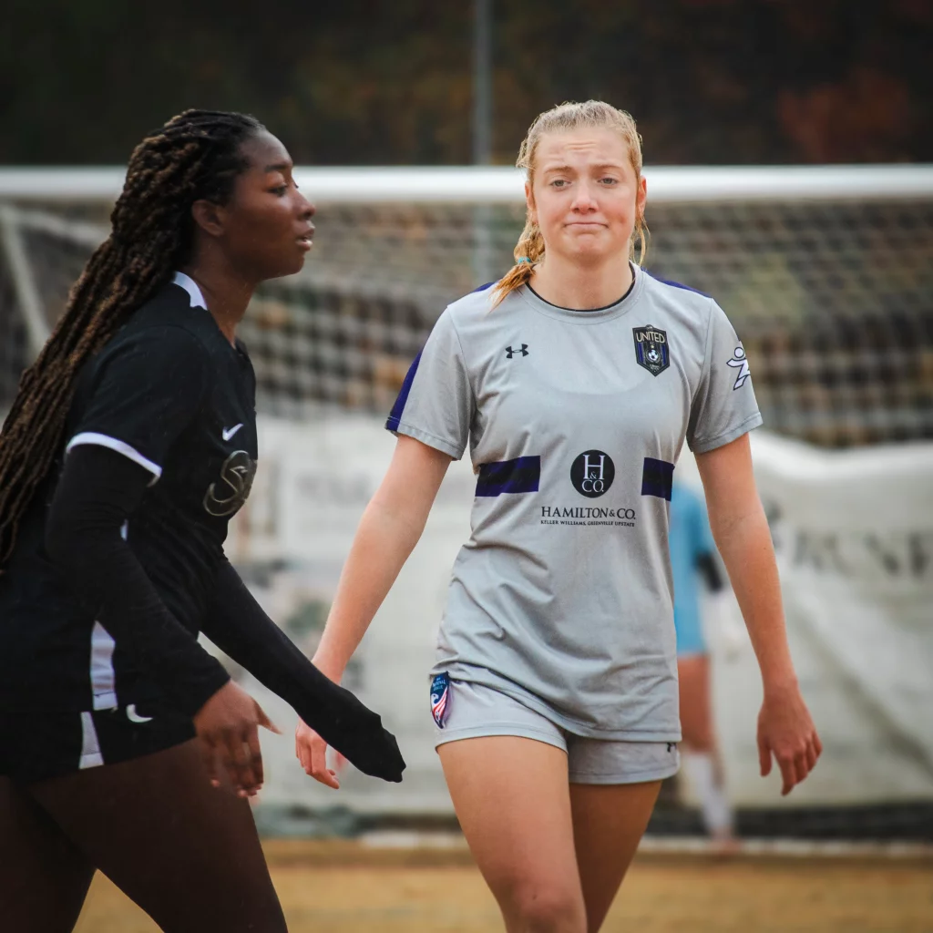 United FC - Furman Region -: Two female soccer players on the field, one in a black uniform and one in a gray uniform, with a goal and net in the background. The player in gray wears a neutral or slightly disappointed expression.