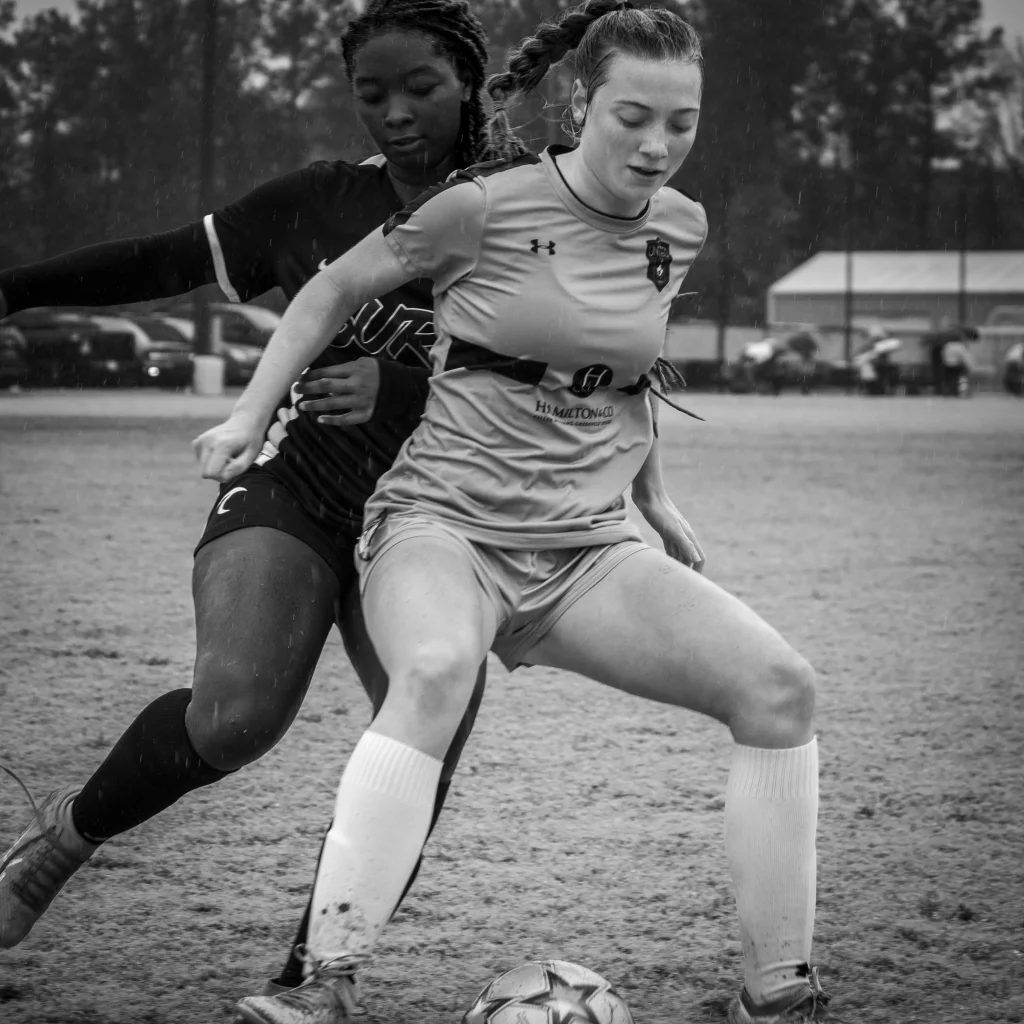 : Black and white photo of two female soccer players competing for the ball on a muddy field, with one player in light-colored uniform in the foreground and another in dark uniform behind her.
