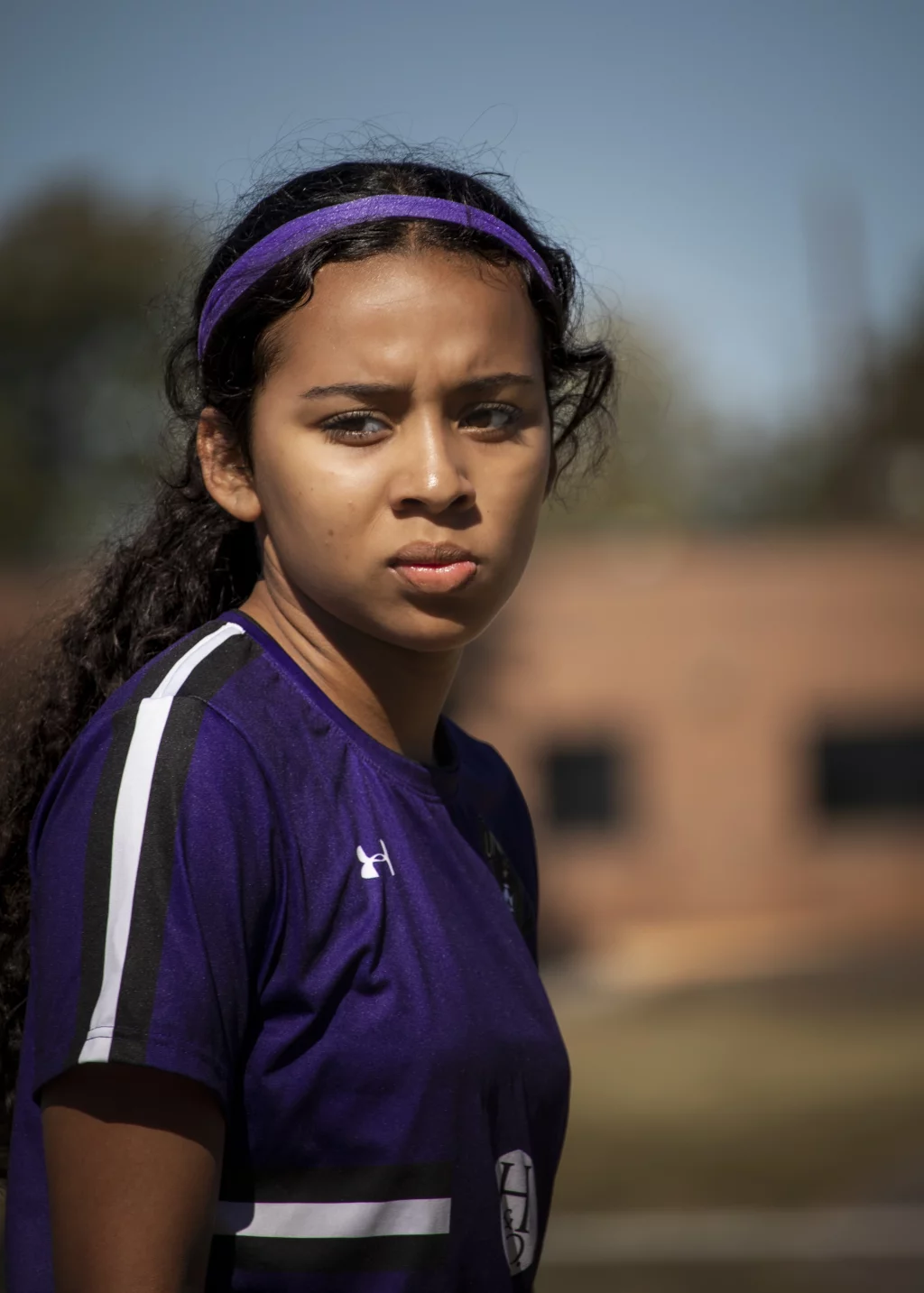 United FC - Furman Region -: A young woman with curly hair, wearing a purple sports jersey and a matching headband, looks seriously towards the camera outdoors with a blurred background.