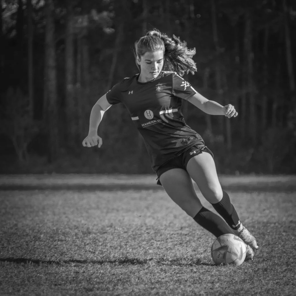 : Black and white photo of a young woman playing soccer, captured mid-stride as she dribbles a ball on a grassy field with trees in the background.