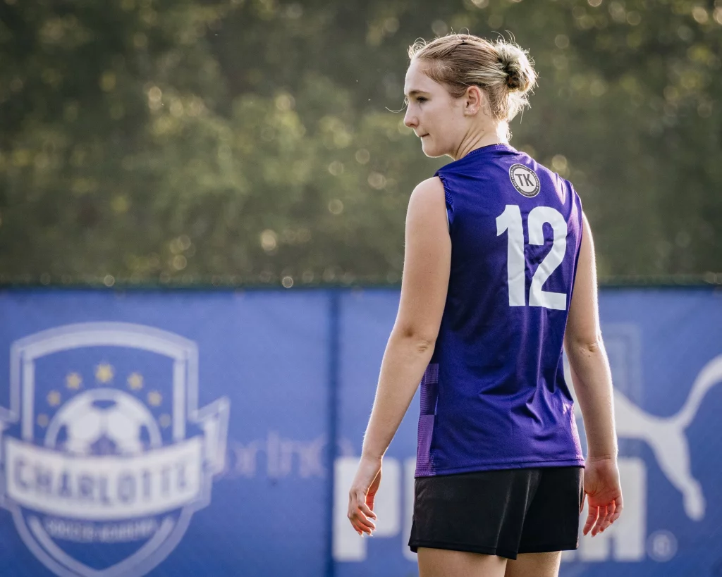 United FC - Furman Region -: A soccer player wearing a purple jersey with the number 12 stands on a field, with a blurred "Charlotte" soccer banner and a Puma logo visible in the background.