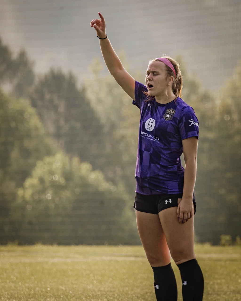 United FC - Furman Region -: A female soccer player in a purple jersey stands on a field with one arm raised and eyes closed, appearing to call out or signal, with trees and soft sunlight in the background.