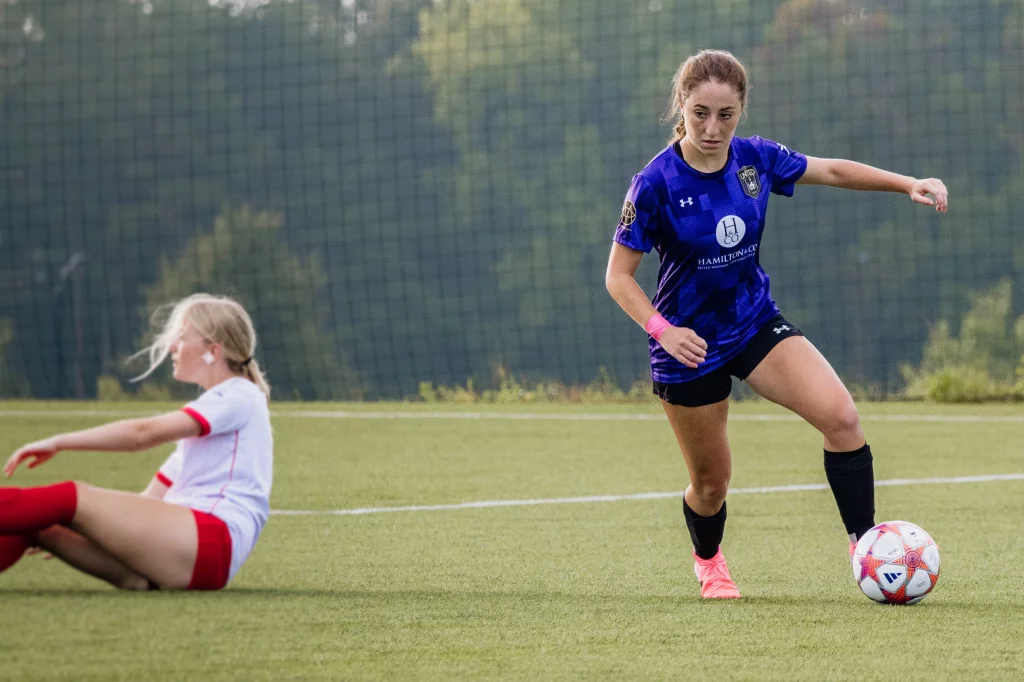 United FC - Furman Region -: Two female soccer players on a field; one in a blue and black uniform dribbles the ball while another in a white and red uniform sits on the ground after a fall.