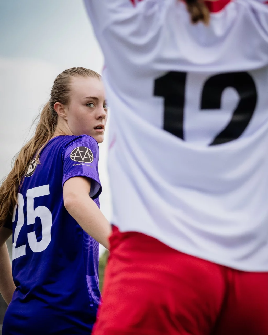 United FC - Furman Region -: A soccer player in a purple jersey with the number 25 looks over her shoulder, while another player in a white jersey with the number 12 and red shorts is in the foreground.