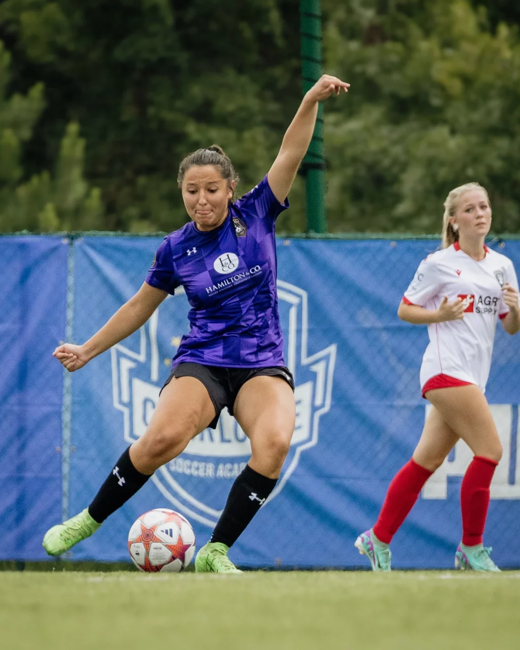 United FC - Furman Region -: Female soccer player in a purple jersey prepares to kick a ball on a field, with another player in a white jersey and red socks in the background.