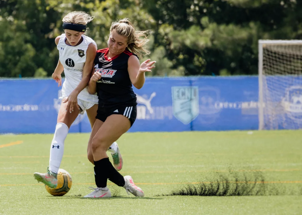 United FC - Furman Region -: Two teenage girls compete for a soccer ball on a field, one in a white uniform and the other in black, with a goal and blue banners in the background.