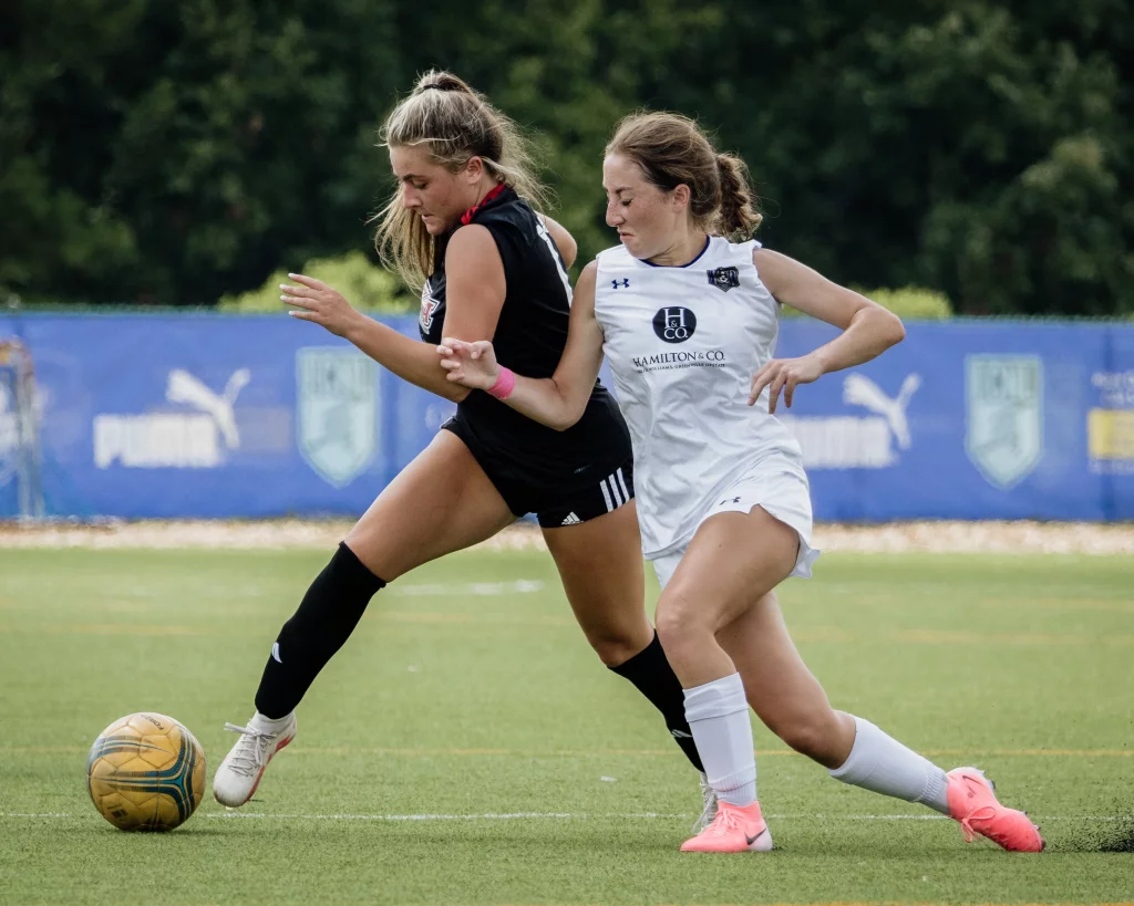 United FC - Furman Region -: Two female soccer players compete for the ball on a field, one in a black uniform and the other in a white uniform, with a blue banner and trees in the background.