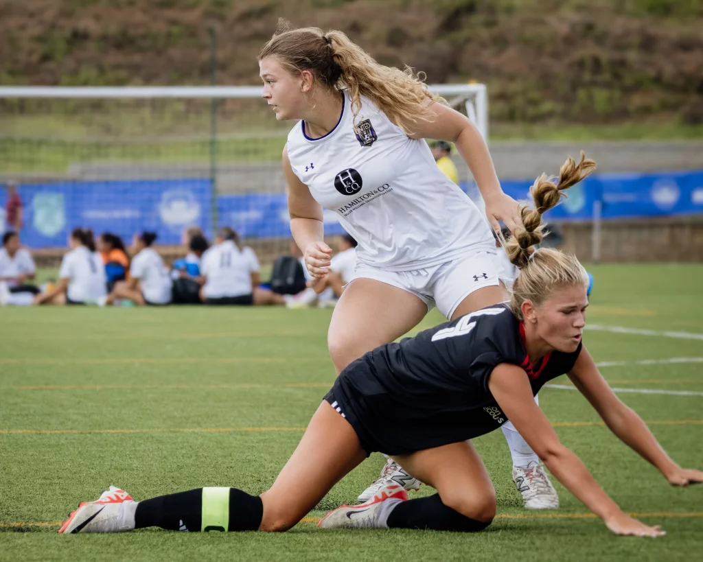 United FC - Furman Region -: Two female soccer players in action; one in a white uniform stands while another in a black uniform is on the ground, with her hands and knees on the turf. A soccer goal and seated players are visible in the background.