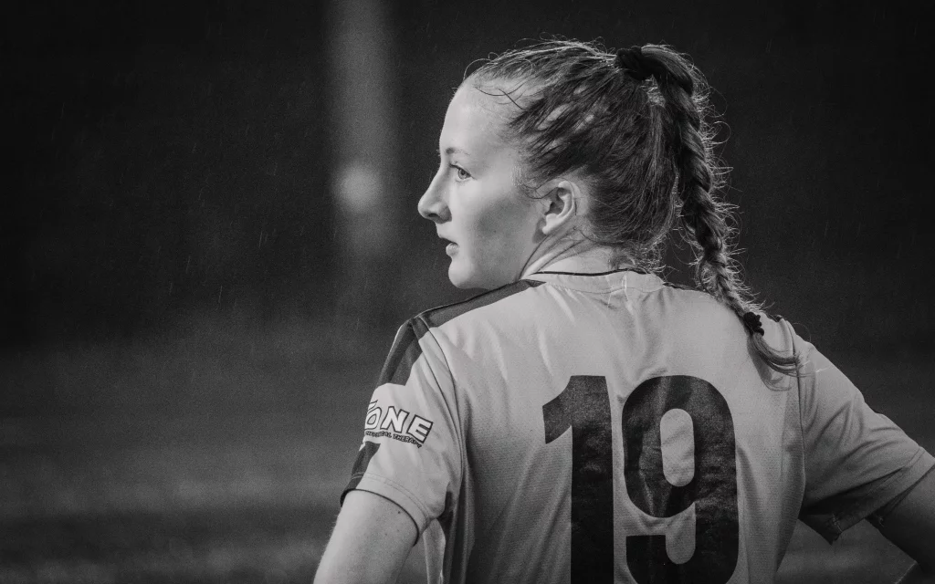 : Black and white photo of a female soccer player with a braid, wearing a jersey with the number 19, looking to her left on a rainy field.