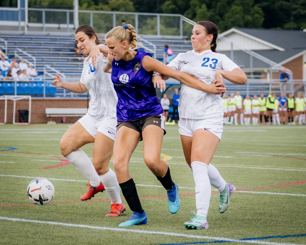 United FC - Furman Region -: Three female soccer players compete for the ball on a field; one in a purple jersey is between two players in white jerseys, with a stadium and spectators in the background.