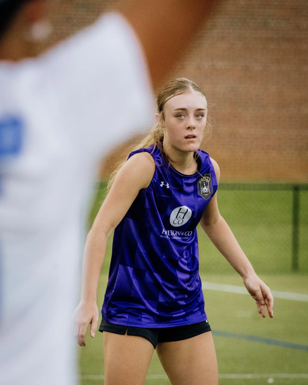 United FC - Furman Region -: A young female soccer player in a purple jersey is focused during a game, standing on a green field with a blurred arm of another player in the foreground.