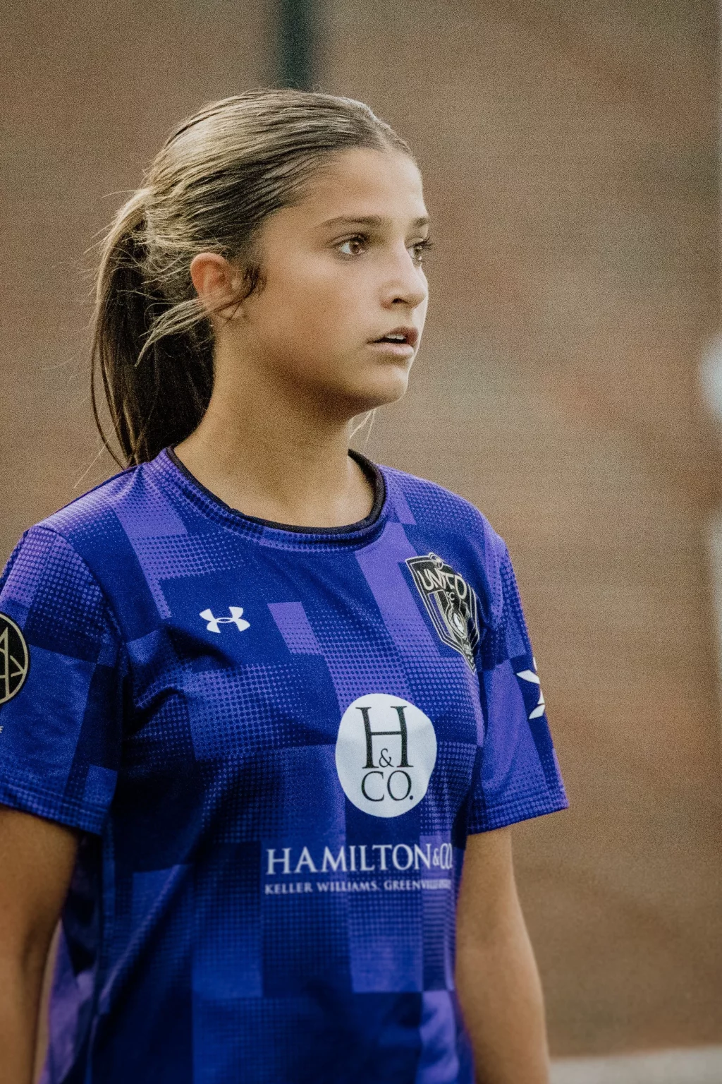 United FC - Furman Region -: Teen girl in a blue soccer jersey with sponsor logos, standing outdoors with a focused expression.