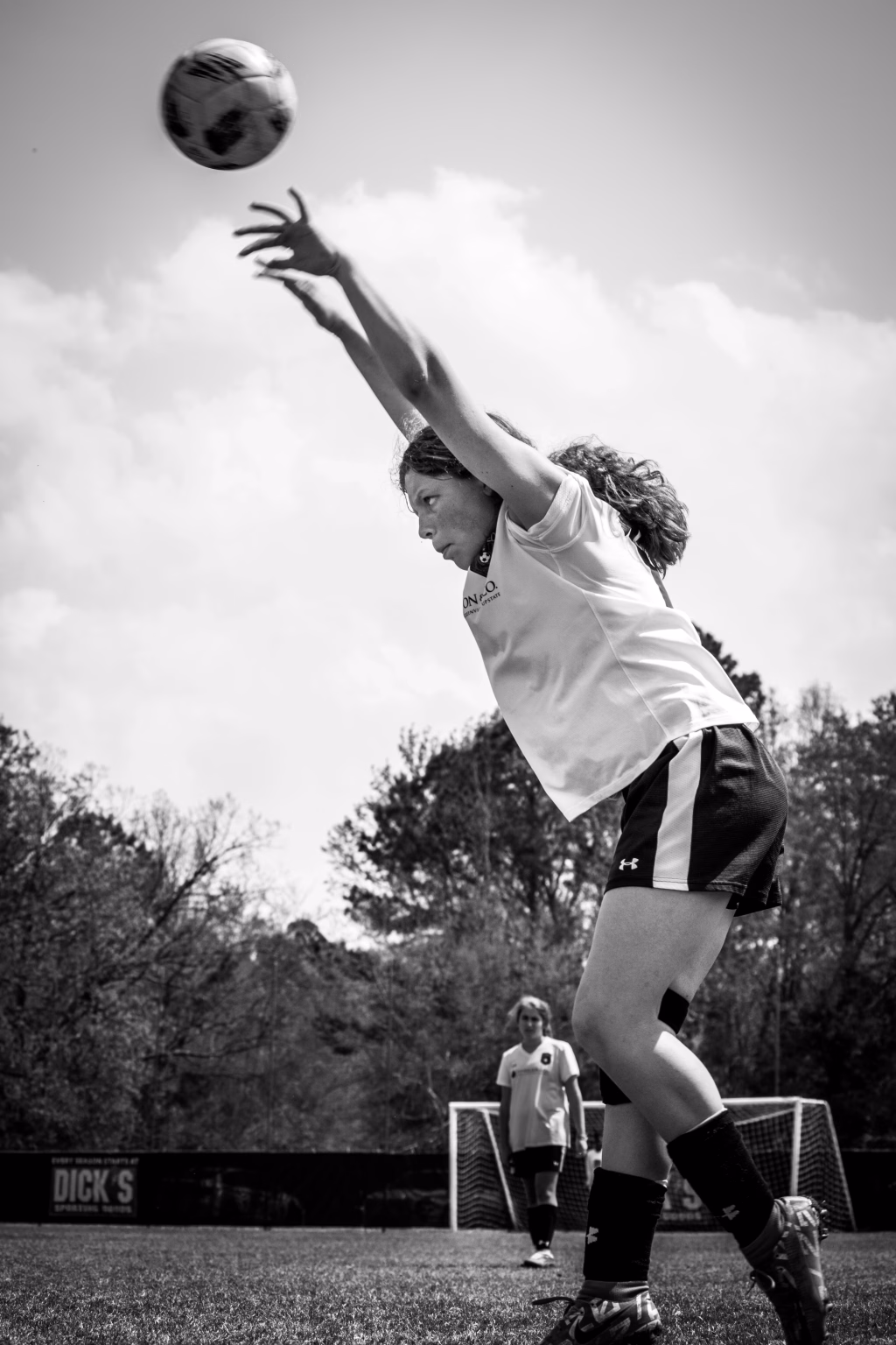 : Black and white photo of a young soccer player in mid-throw, launching a soccer ball overhead during a game, with another player and a goal visible in the background.