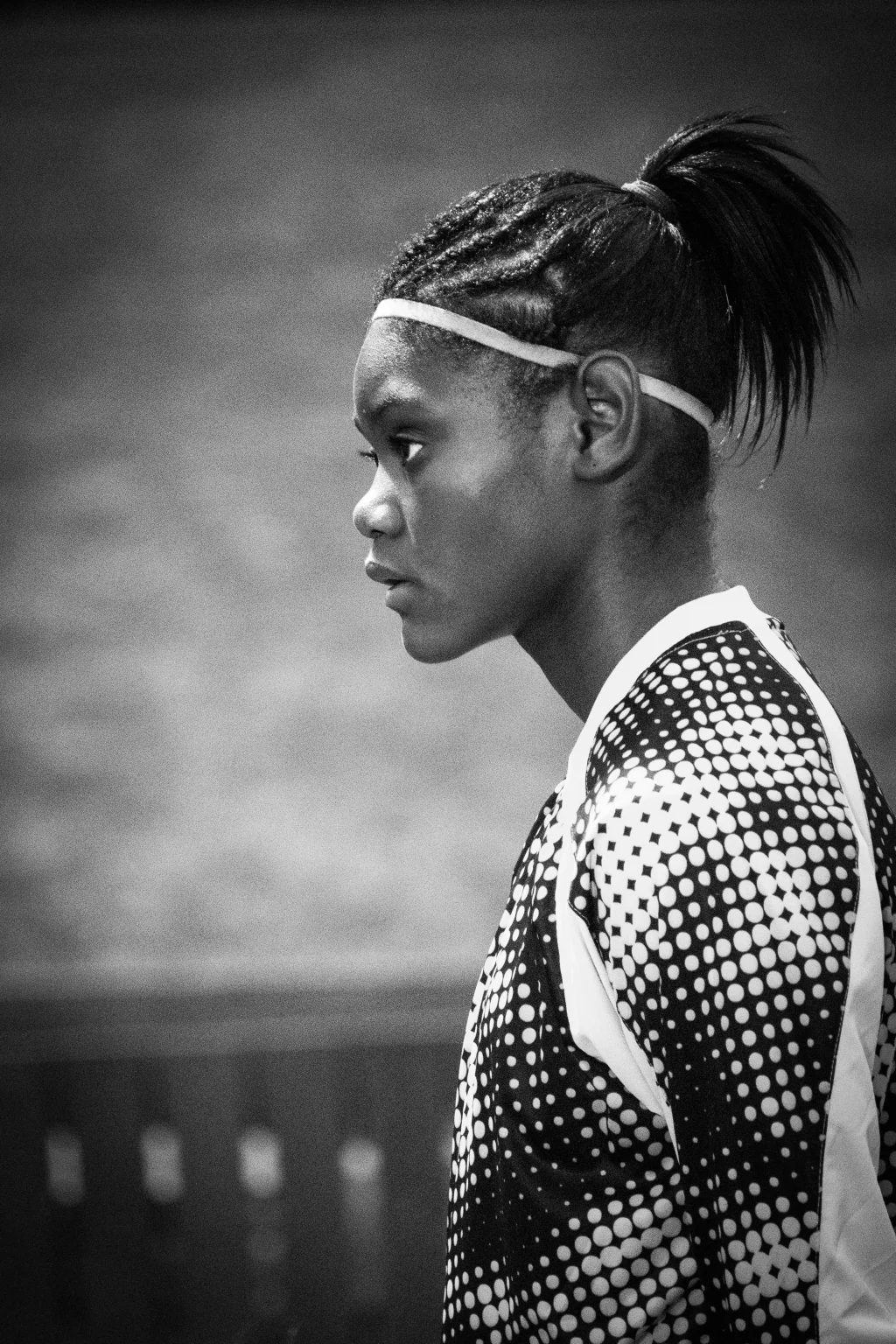 : Black and white photo of a focused young athlete in profile, wearing a patterned sports jersey and a headband, with braided hair pulled back into a ponytail.
