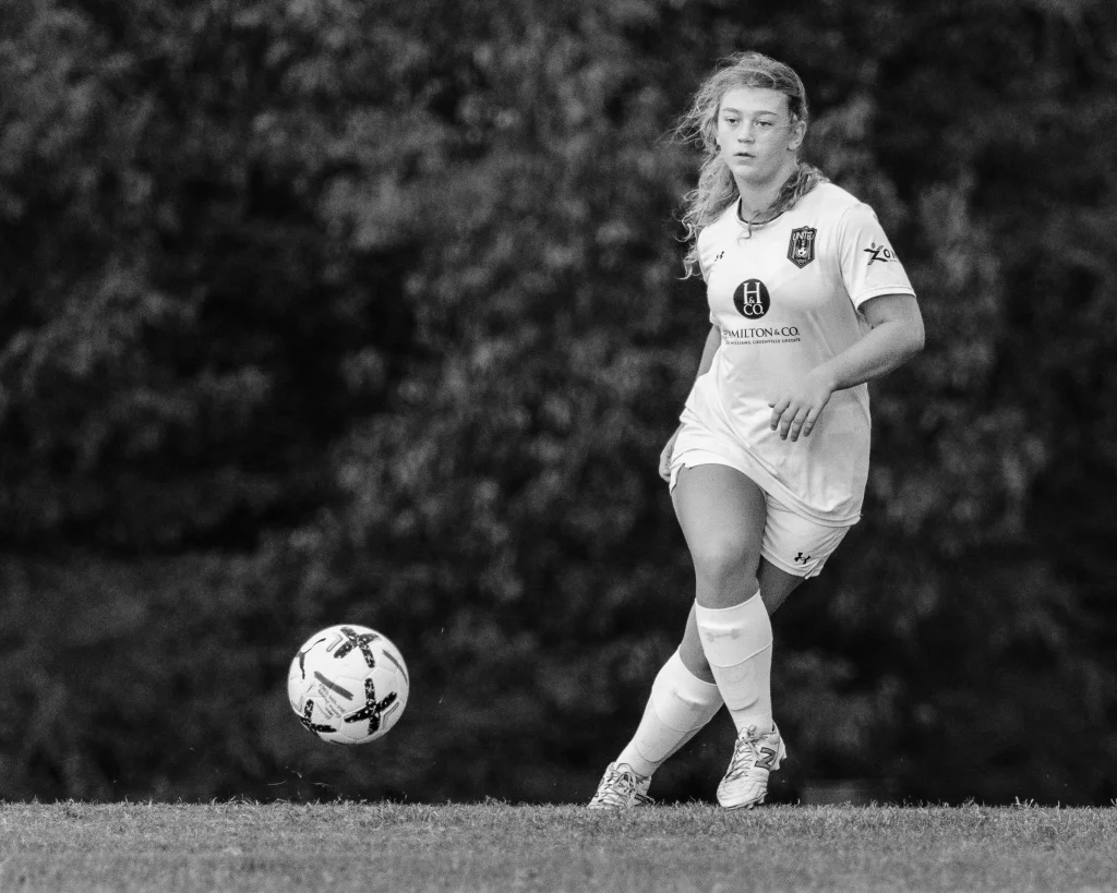 : Black and white photo of a female soccer player in a white uniform running toward a soccer ball on a grassy field, with blurred trees in the background.
