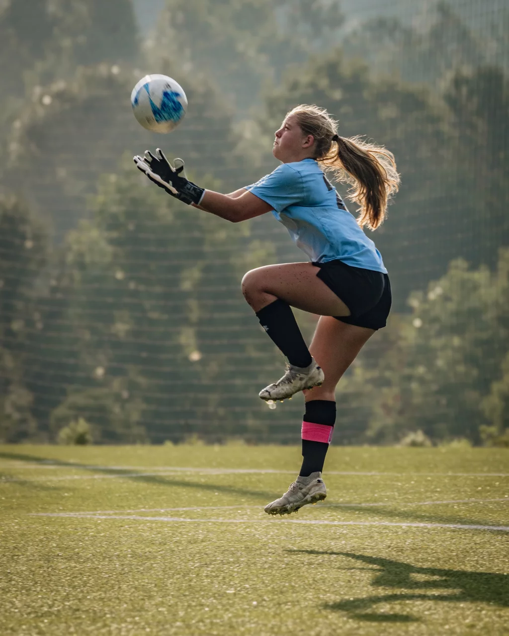 United FC - Furman Region -: Soccer goalkeeper in a blue jersey and gloves jumps to catch a ball, with one knee raised, on a grassy field.
