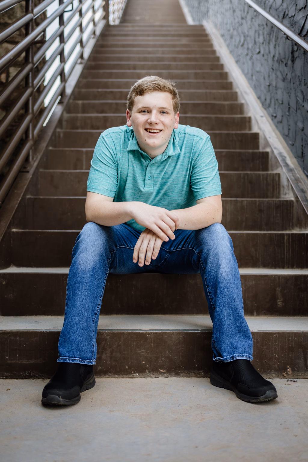 Wesley's senior photo taken at BridgeWay Station, Simpsonville, SC: Young man sitting on outdoor concrete stairs, smiling at the camera. He is wearing a light green polo shirt, blue jeans, and black shoes. Metal railings are visible on both sides of the stairs.