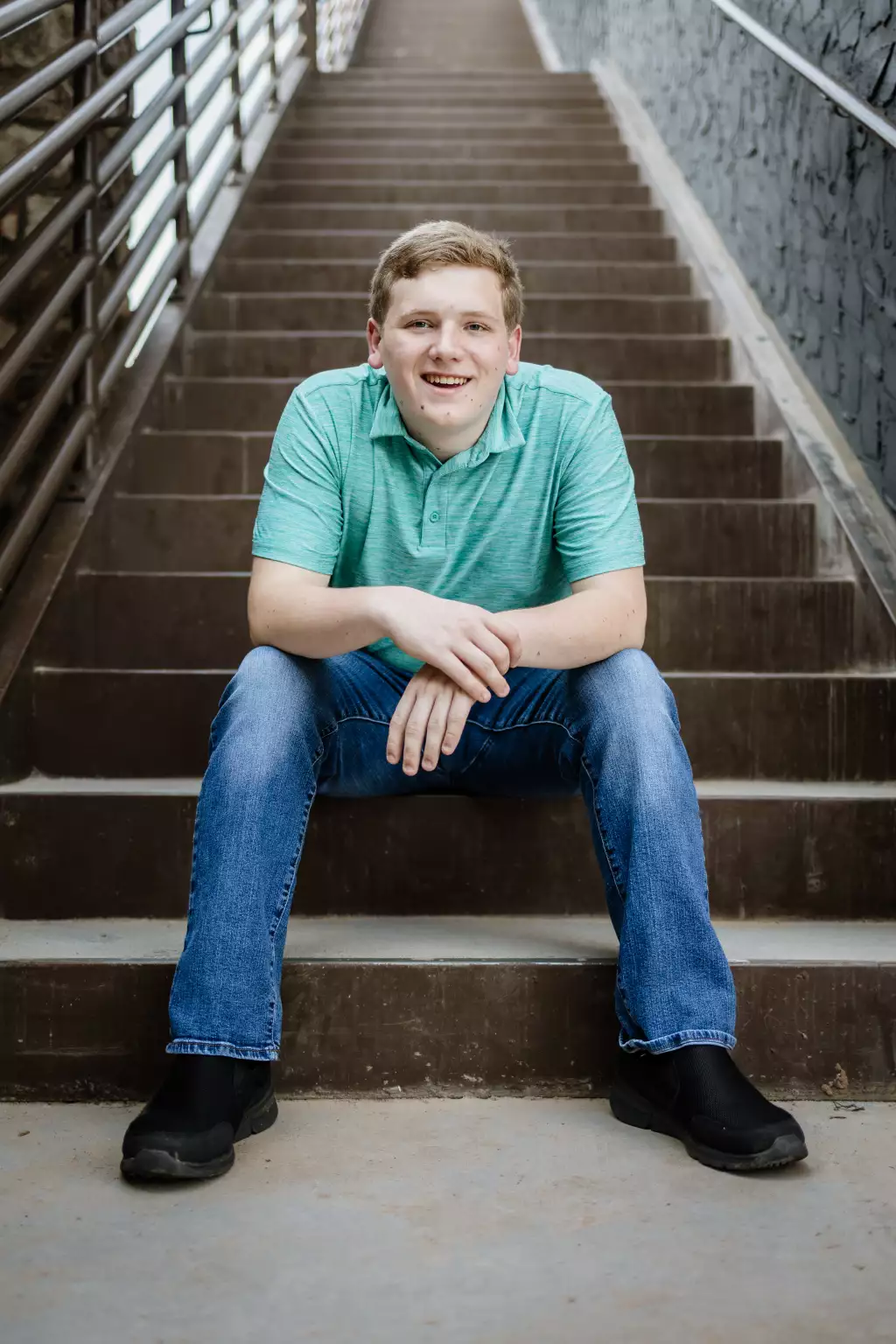 : Young man sitting on outdoor concrete stairs, smiling at the camera. He is wearing a light green polo shirt, blue jeans, and black shoes. Metal railings are visible on both sides of the stairs.