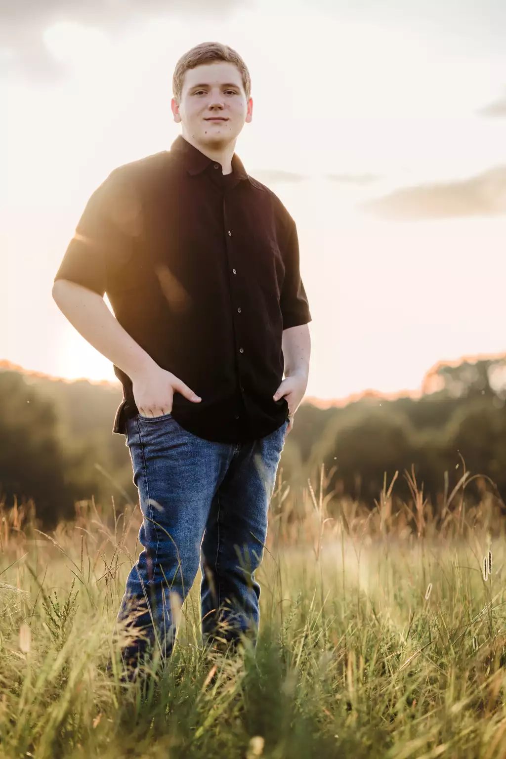 : Young man standing in a sunlit grassy field at sunset, wearing a black button-up shirt and blue jeans, with hands in pockets and a slight smile.