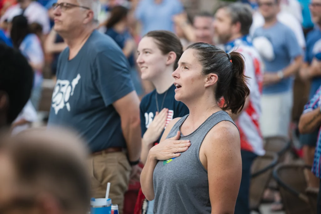 : A group of people standing with their right hands over their hearts, appearing to sing or recite, likely during a national anthem at a sporting event.