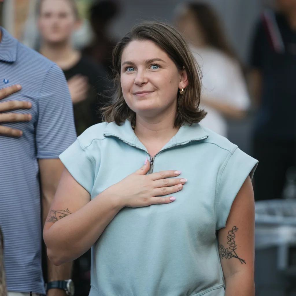 : Woman standing with her hand over her heart, looking upward, surrounded by other people doing the same gesture in an outdoor setting.