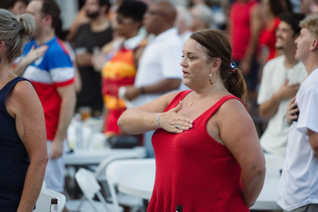 : A woman in a red top stands with her hand over her heart among a crowd, likely during a national anthem or moment of respect. White folding chairs and tables are visible in the background.