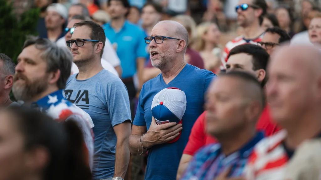 : A group of people standing close together, with one man in the center holding a cap to his chest and singing, appearing solemn and attentive, likely during a national anthem at a sporting event.