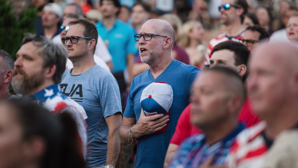 : A group of people standing close together, with one man in the center holding a cap to his chest and singing, appearing solemn and attentive, likely during a national anthem at a sporting event.