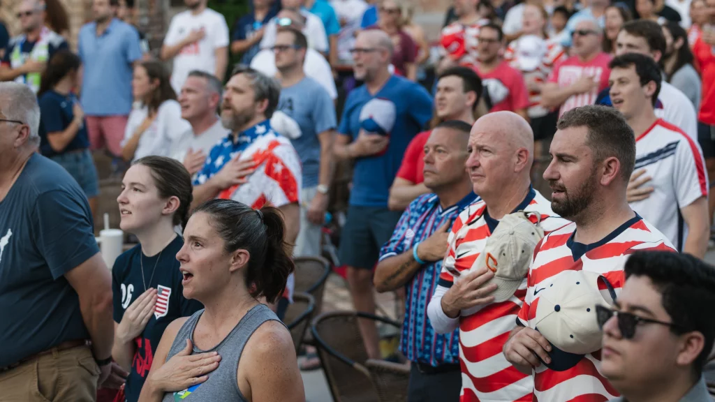 : A large group of people, many wearing red, white, and blue, stand with hands over their hearts, appearing to sing or recite, likely during a national anthem at a sporting event.