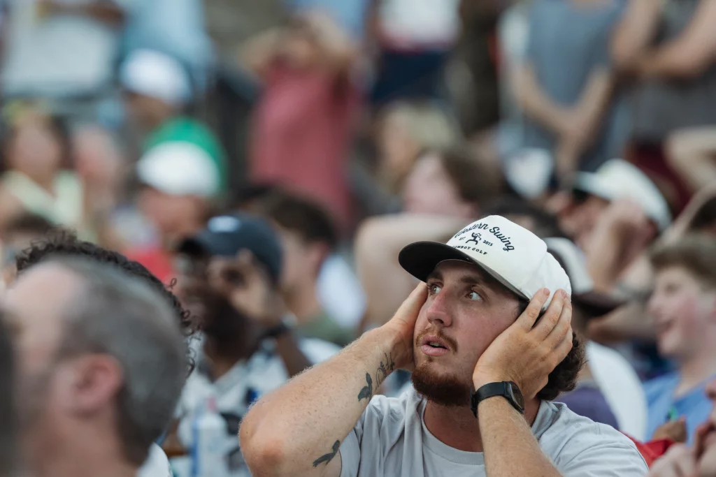 : A man in a crowd covers his ears and looks upward with a worried expression, surrounded by other spectators, some also showing concerned body language.