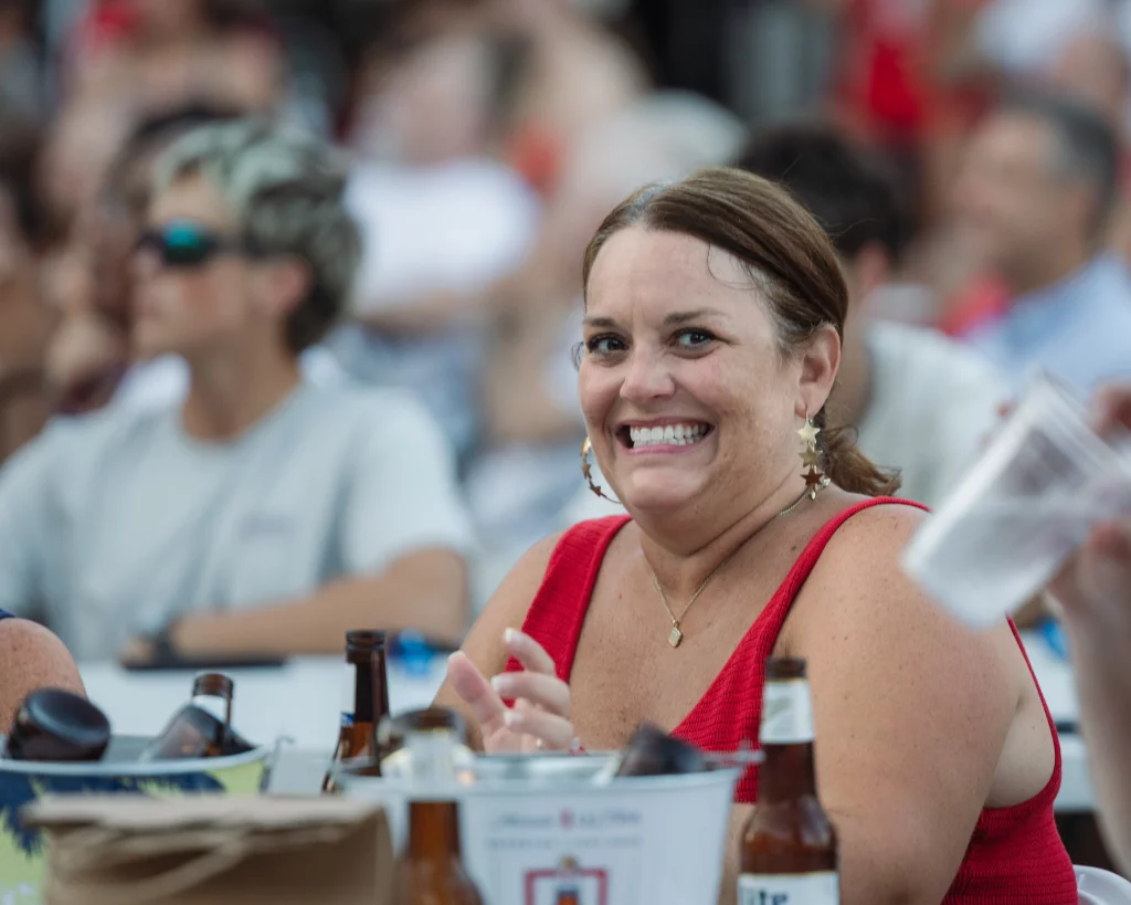 : Woman in a red tank top smiling at an outdoor event, with drinks and bottles on the table and a crowd in the blurred background.