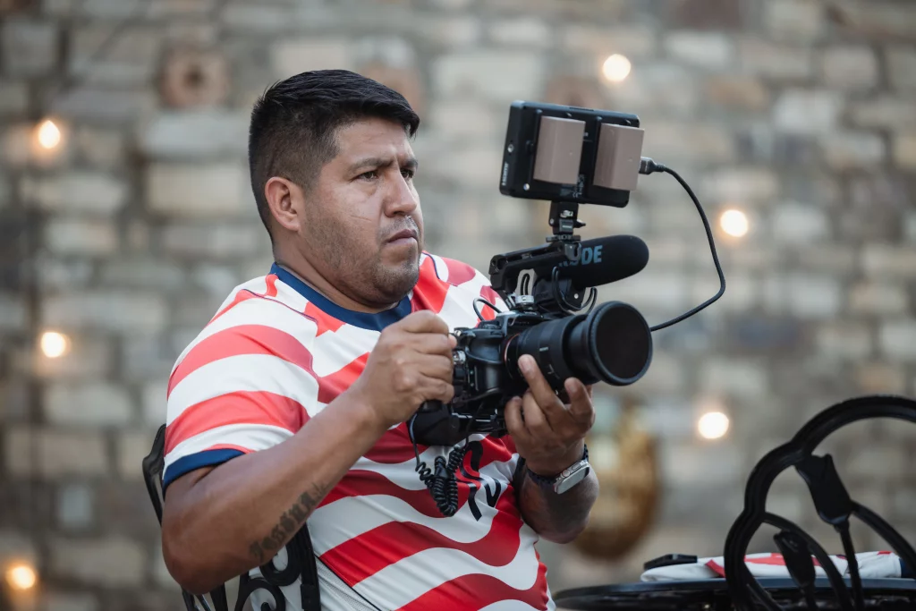 : Man in a red and white striped shirt holding a professional video camera with a microphone and monitor, seated in front of a blurred stone wall with string lights.