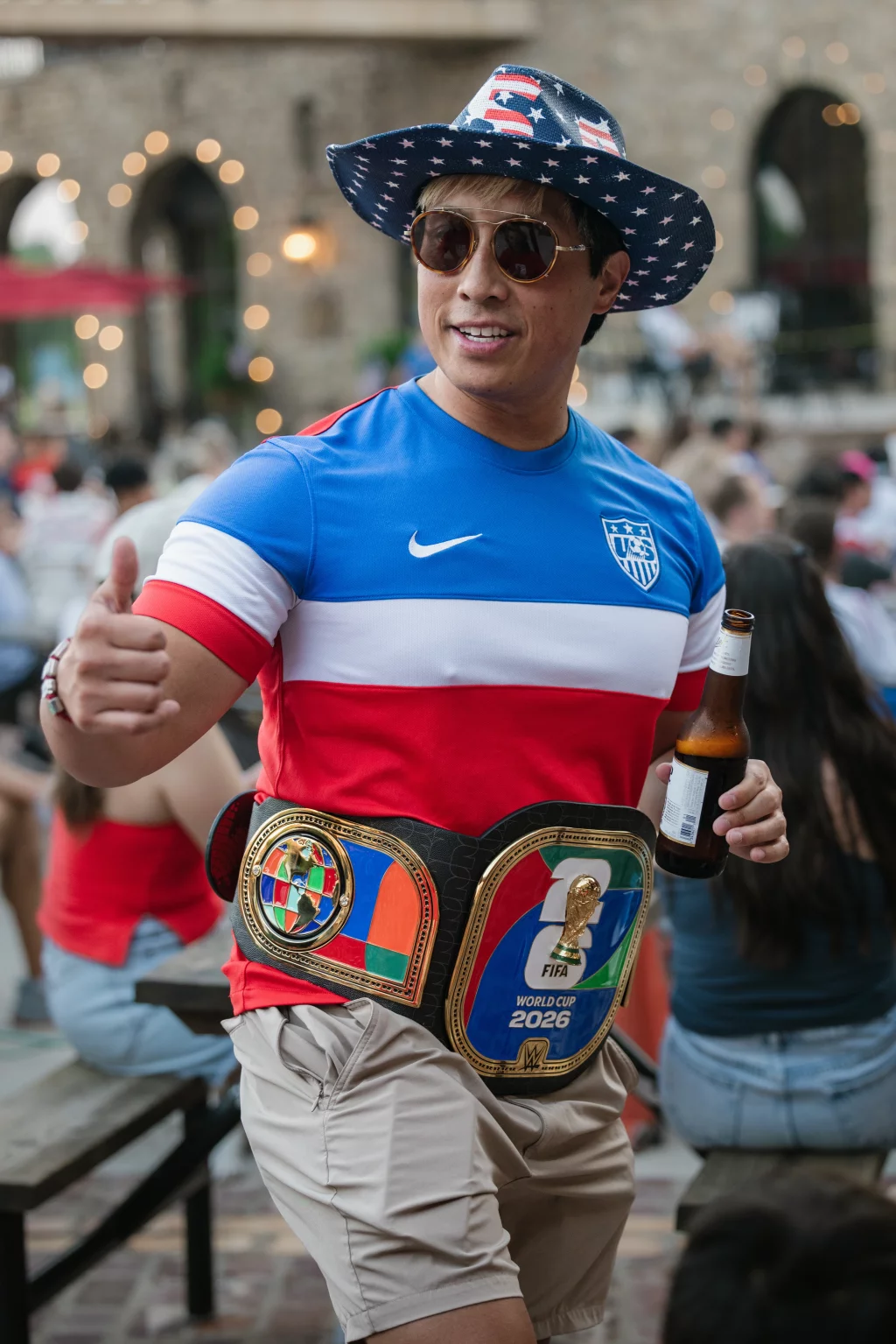: Man wearing a USA soccer jersey, star-spangled hat, and sunglasses, holding a beer and giving a thumbs up, with a large World Cup 2026-themed championship belt around his waist.