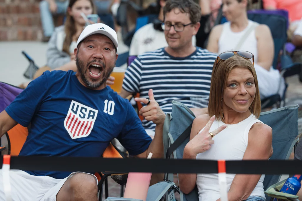 : A man in a blue USA soccer shirt excitedly points at a woman beside him who is smiling and making a peace sign, both sitting in outdoor chairs among a crowd.