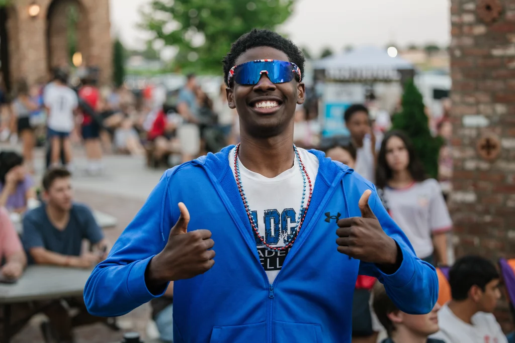 : Smiling person wearing reflective sunglasses, a blue jacket, and beaded necklaces gives two thumbs up in a crowded outdoor setting.