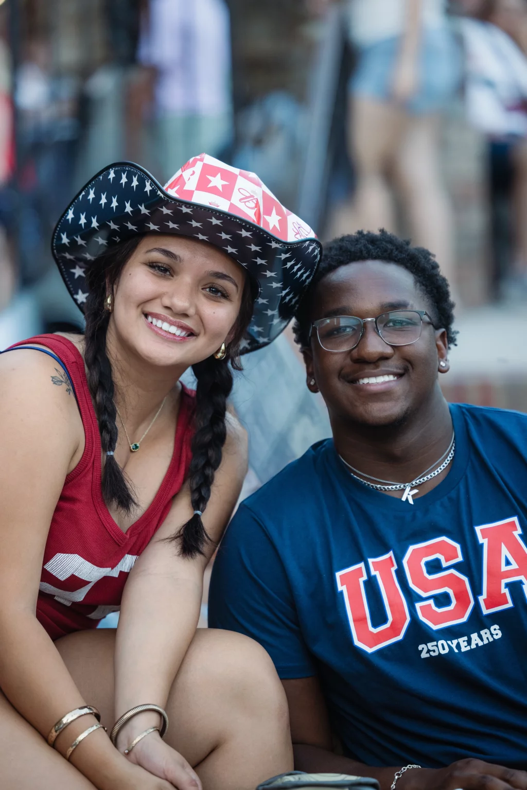 : Two people smiling at the camera; one wears a star-patterned cowboy hat and red tank top, the other wears glasses and a blue "USA 250 YEARS" shirt.