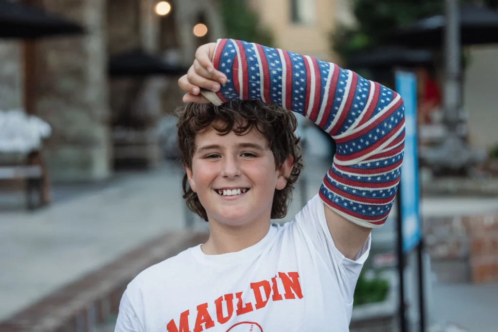 : Smiling boy wearing a white "Mauldin" t-shirt, with a red, white, and blue American flag-patterned arm cast, raising his injured arm above his head.