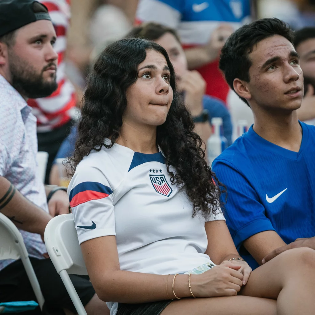 : A young woman in a USA soccer jersey and a young man in a blue shirt sit closely together, watching something intently with anxious expressions among a crowd.