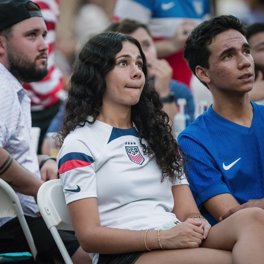 : A young woman in a USA soccer jersey and a young man in a blue shirt sit closely together, watching something intently with anxious expressions among a crowd.