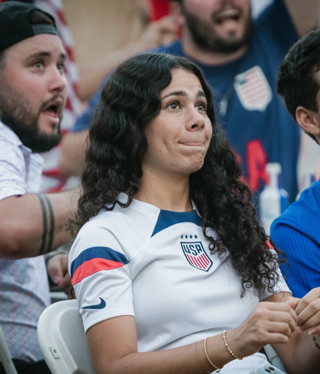: A group of young fans watch a sporting event with anxious expressions, including a woman in a white USA soccer jersey and a man in a blue jersey, seated among a crowd.