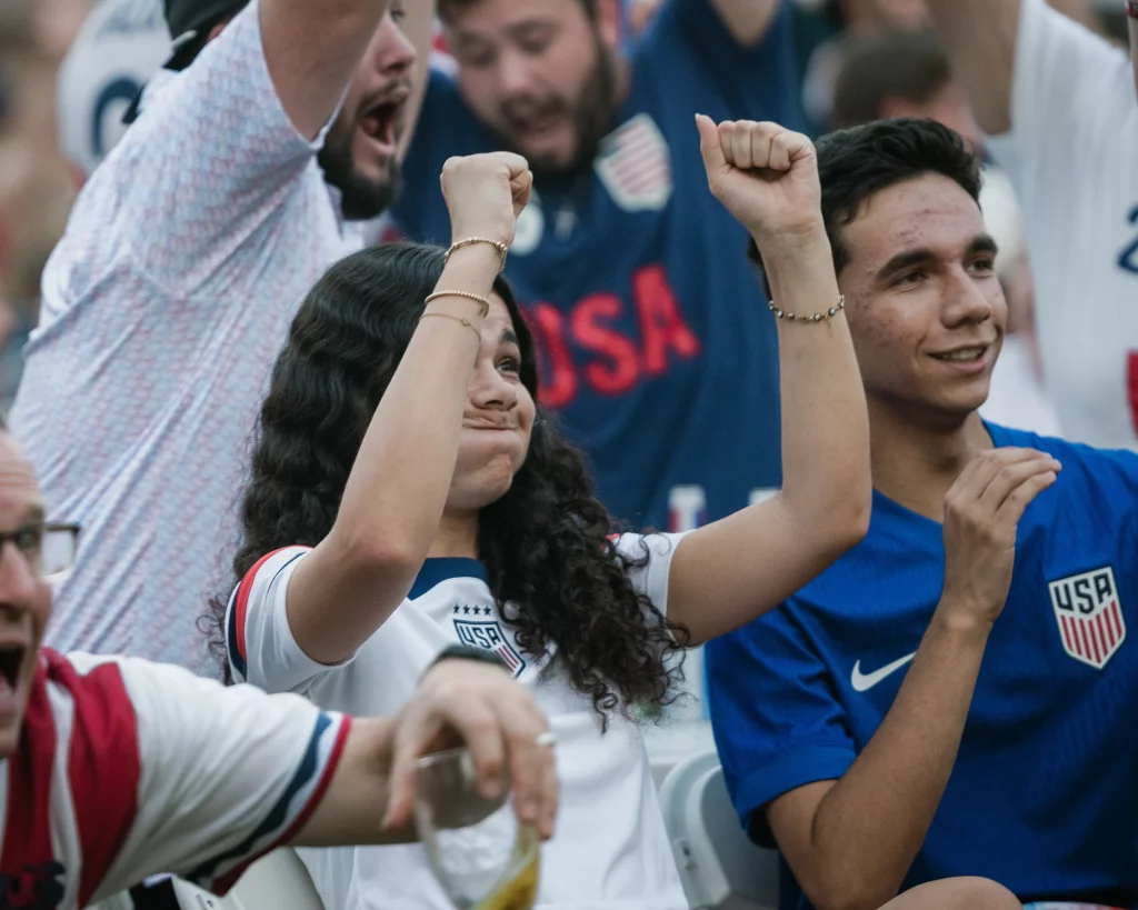 : Fans wearing USA soccer jerseys cheer with raised arms, showing excitement and celebration in a crowded setting.