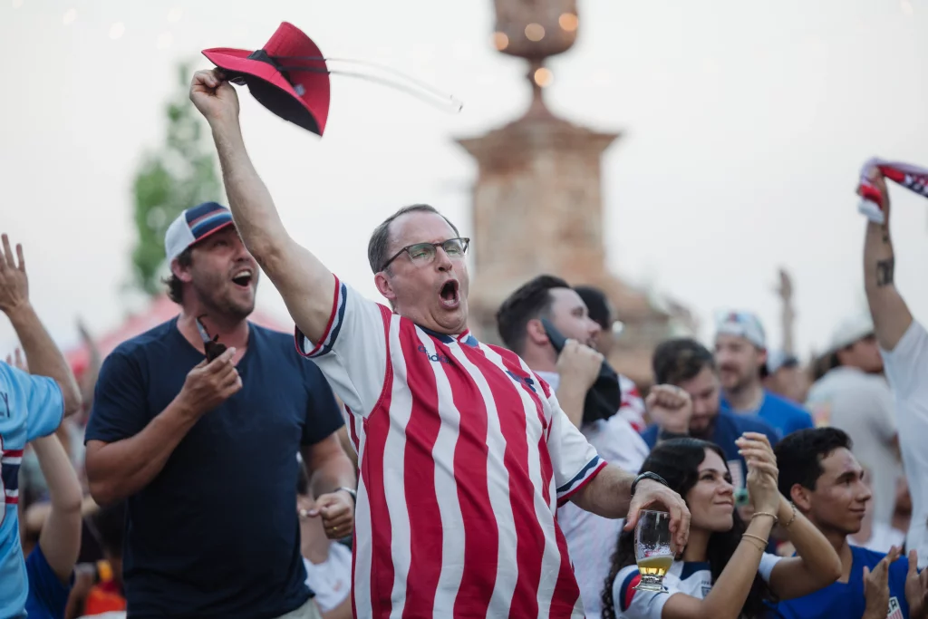 : A group of enthusiastic soccer fans cheer outdoors, with a man in a red and white striped jersey raising a hat and holding a drink, surrounded by others clapping and celebrating.