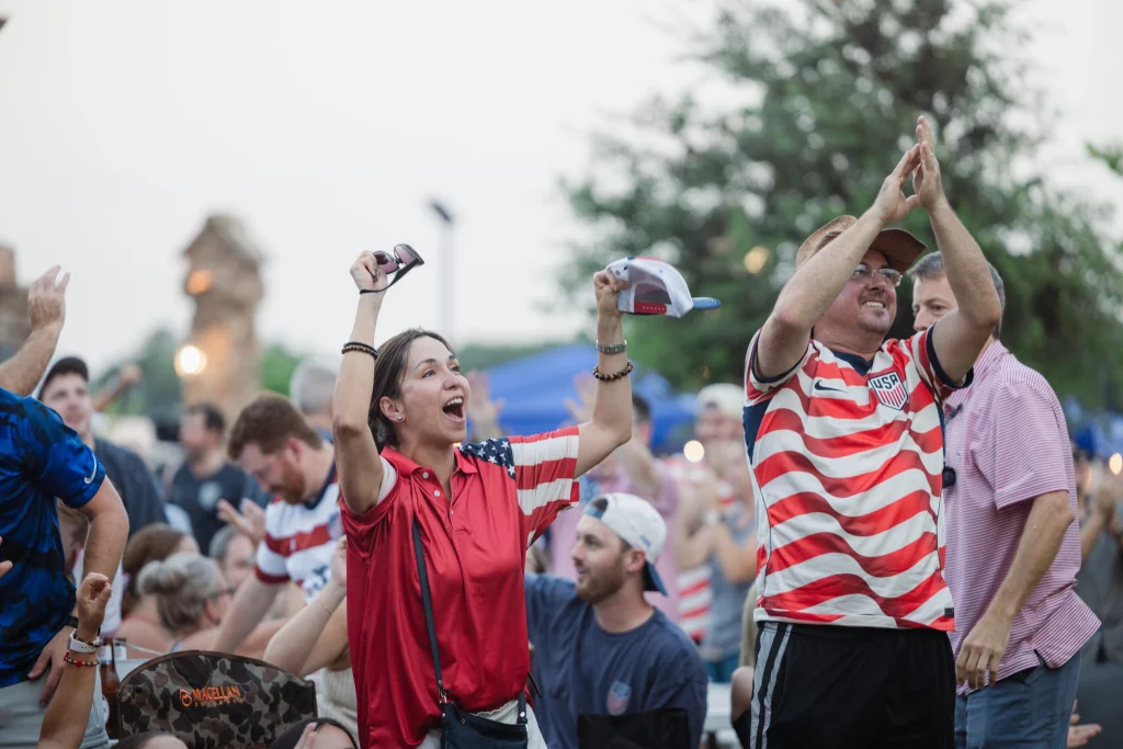 : A crowd of people outdoors enthusiastically cheering, with two people in the foreground wearing USA-themed clothing and raising their arms in celebration.
