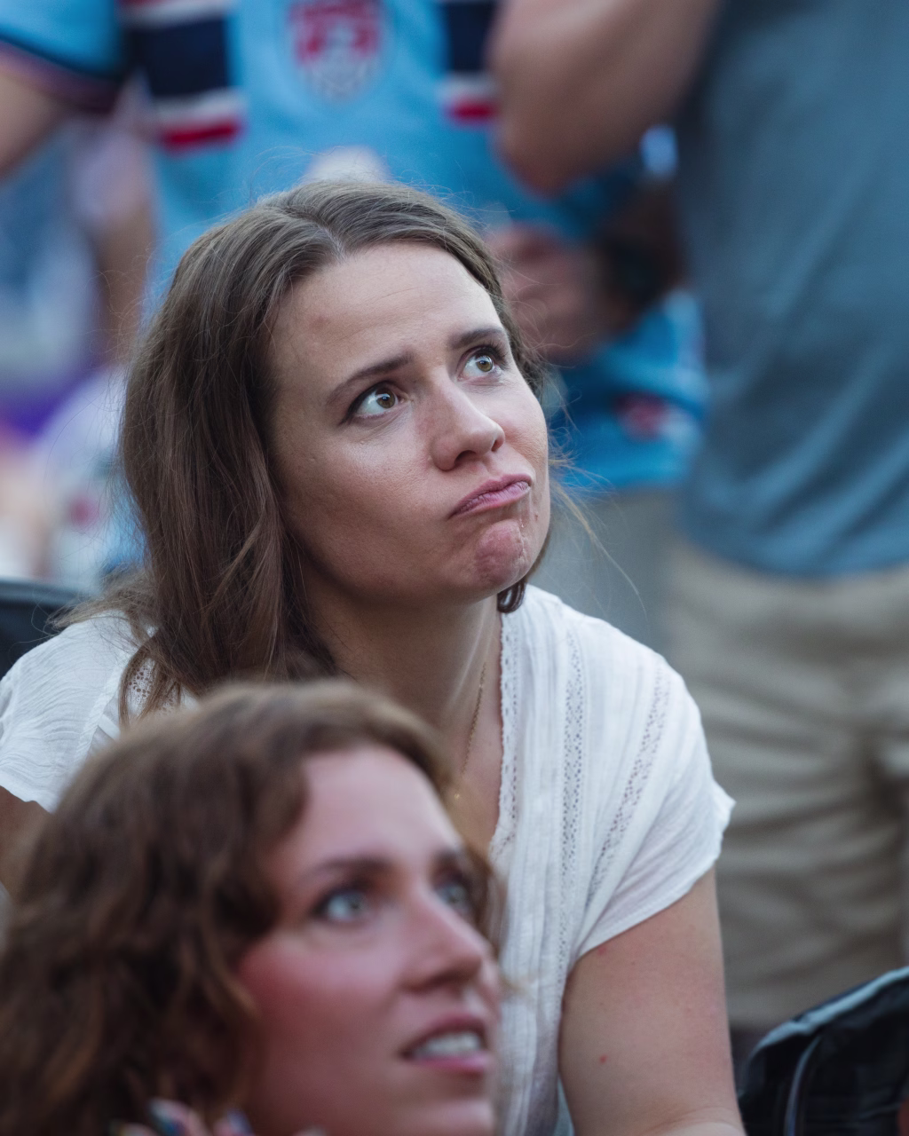 : A woman with long brown hair in a white shirt looks upward with a disappointed or concerned expression. Another person is visible in the foreground, slightly out of focus. The background shows people wearing blue sports jerseys.