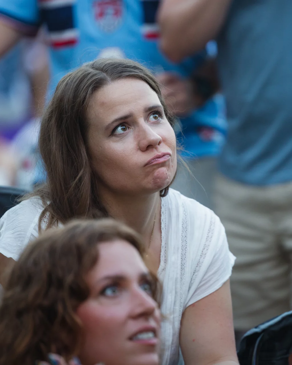 : A woman with long brown hair in a white shirt looks upward with a disappointed or concerned expression. Another person is visible in the foreground, slightly out of focus. The background shows people wearing blue sports jerseys.
