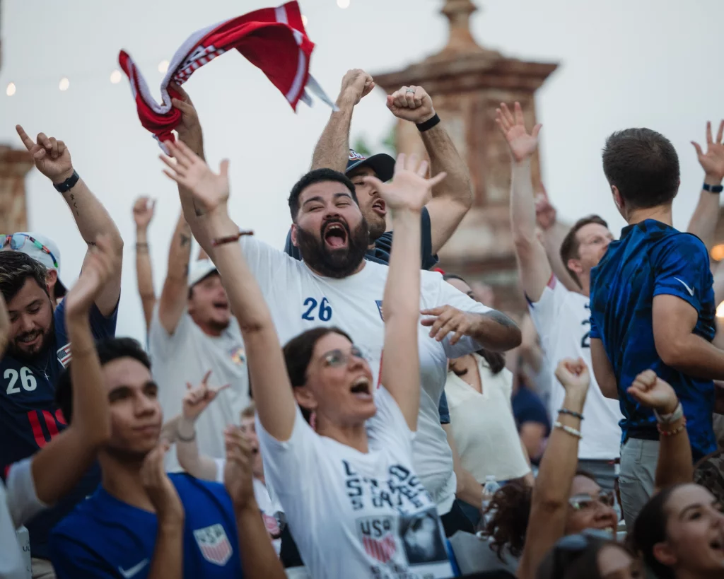 : A crowd of enthusiastic soccer fans cheer with raised arms, some wearing USA jerseys and waving a red and white scarf, celebrating outdoors.