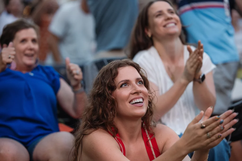 : Three women in a crowd are smiling, clapping, and giving thumbs up, appearing excited and happy while watching an event.