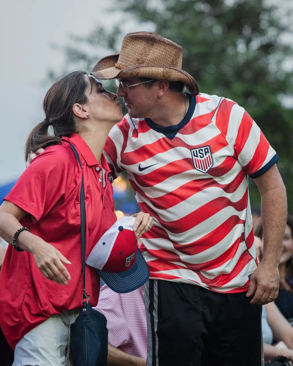 : A man and woman wearing USA soccer fan gear share a kiss outdoors; the man wears a cowboy hat and a red-and-white striped USA jersey, while the woman holds a USA cap and wears a red shirt.