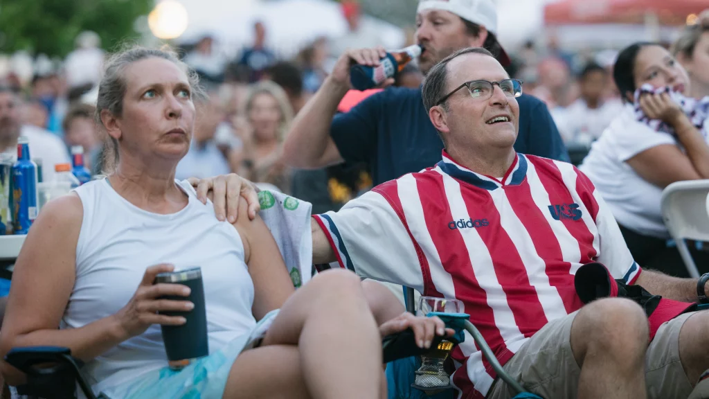 : A woman and a man sit closely together in a crowd, both watching something intently off camera. The man wears a red and white striped USA soccer jersey and glasses, while the woman holds a drink. The atmosphere suggests they are at a public viewing event, possibly for a sports match.