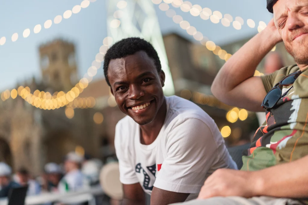 : Two men sit outdoors in a festive setting with string lights in the background; one man smiles directly at the camera while the other looks relaxed with his eyes closed.